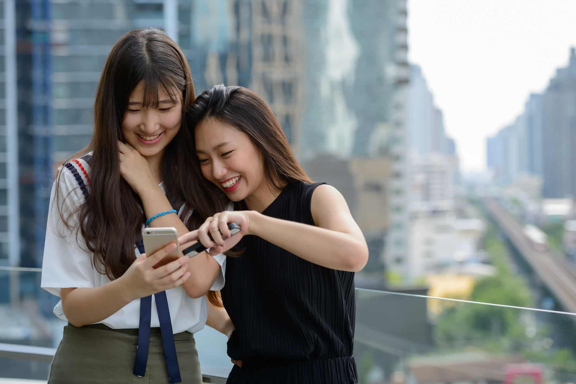 Two happy young beautiful Asian teenage girls using phone together against view of the city