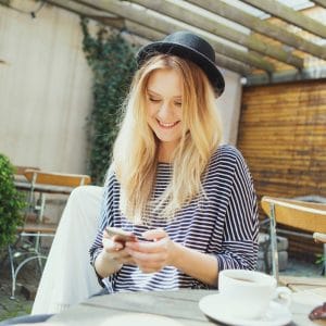 Smiling young woman wearing hat using phone while having coffee at cafe