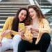 Two women smiling and having fun while using mobile phone sitting on stairs outdoors.